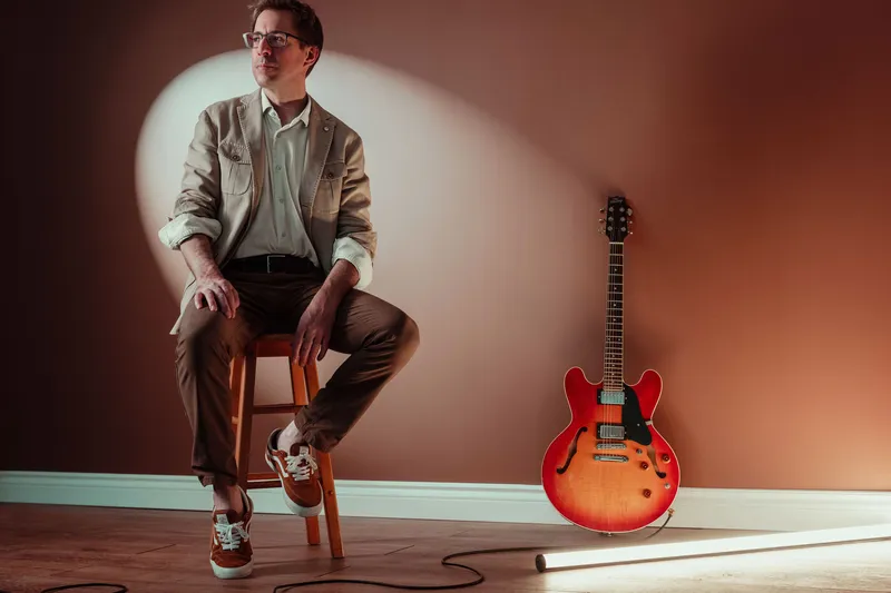Homme à lunettes assis sur un tabouret, éclairé par un projecteur, près d'une guitare électrique rouge appuyée contre un mur orangé.